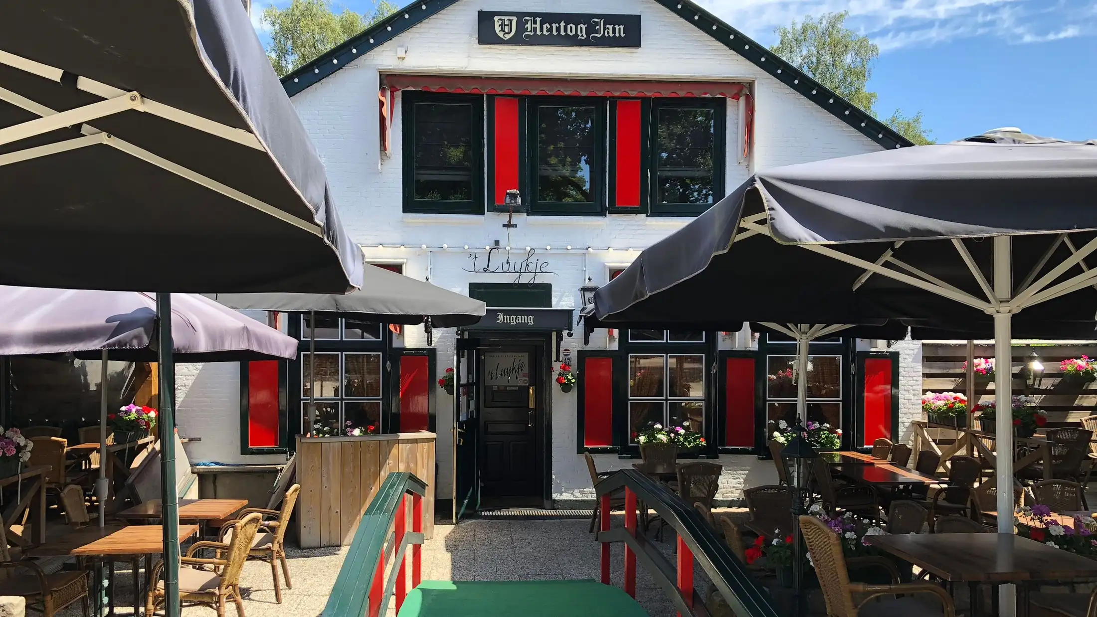 De witte gevel van 't Luykje met rode luiken, het terras met parasols en de groene loopbrug in de zon